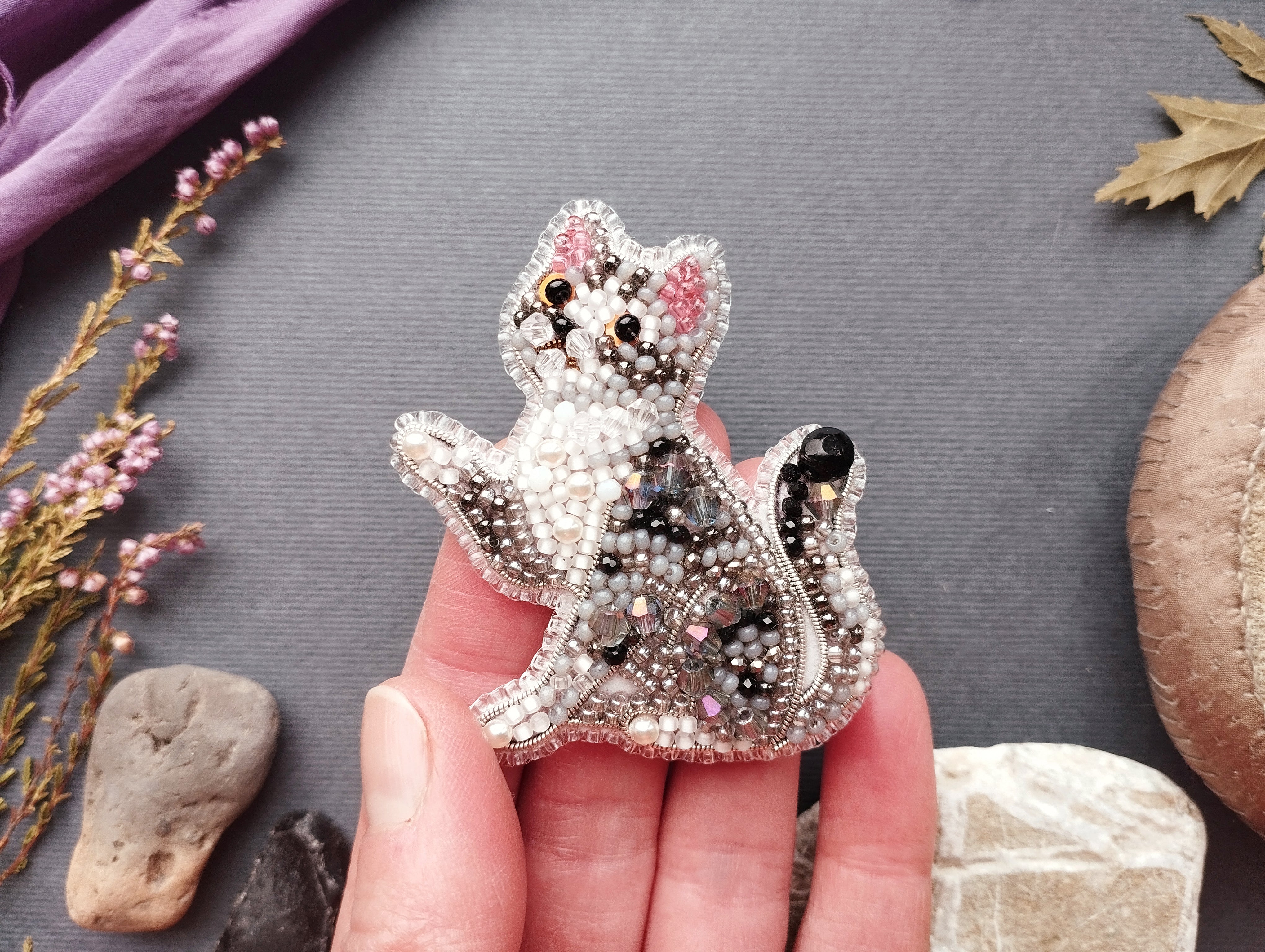 A hand holding a decorative beaded cat brooch against a gray background.